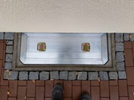 Aluminium Lichtschachtabdeckung mit zwei Halterungen auf einem gepflasterten Boden.