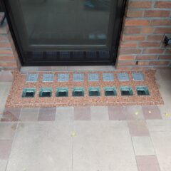 Lichtschachtabdeckung mit mehreren Gitteröffnungen und Steinverkleidung vor einem Fenster.