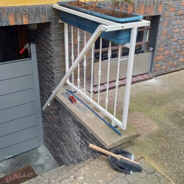 Graue Zugangstür mit einer Treppe und einem Blumenkasten darüber, der an einem Geländer befestigt ist.