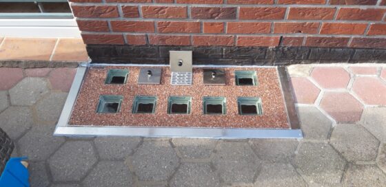 Entwässerungssystem mit mehreren Glassteinen und einem Abfluss in der Mitte auf einem gepflasterten Boden.