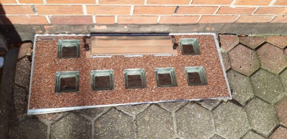 Abdeckung mit Glaselementen auf einem gepflasterten Boden, umgeben von Ziegelmauerwerk.