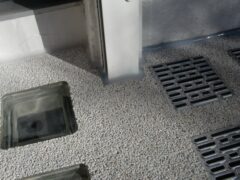 Lichtschachtabdeckungen aus Metall und Gitterroste auf einer Oberfläche mit Kieselsteinen.