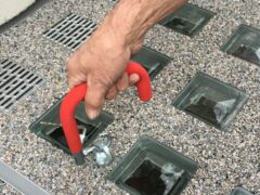 Eine Hand mit einem roten Griff, die eine Lichtschachtabdeckung anhebt, um darunter liegende Glassteine zu zeigen.