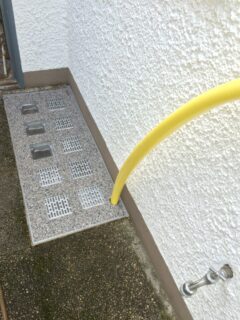 Lichtschachtabdeckung mit mehreren Gitteröffnungen und einem gelben Schlauch, der daran angeschlossen ist.