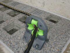 Graue Lichtschachtabdeckung mit grünen Halterungen und einem schwarzen Kabel auf einem Kiesbett.