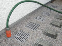 Lichtschachtabdeckungen aus grauem Material mit einem grünen Gartenschlauch und einem orangefarbenen Anschluss.