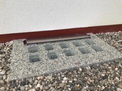 Lichtschachtabdeckung aus Stein mit integrierten Glassteinen auf einem Kiesbett.
