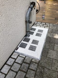 Lichtschachtabdeckung mit mehreren Gitterelementen und einem Entwässerungsrohr an einer Wand.
