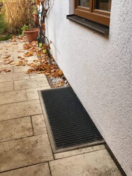 Gitterrost zur Wasserableitung an der Wand eines Gebäudes mit herabgefallenen Blättern im Vordergrund.
